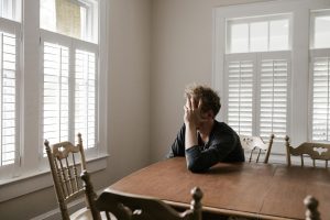 A frustrated man sits alone at a dining table with his hand covering his face, reflecting the anxiety and stress homeowners feel when their house is not selling.
