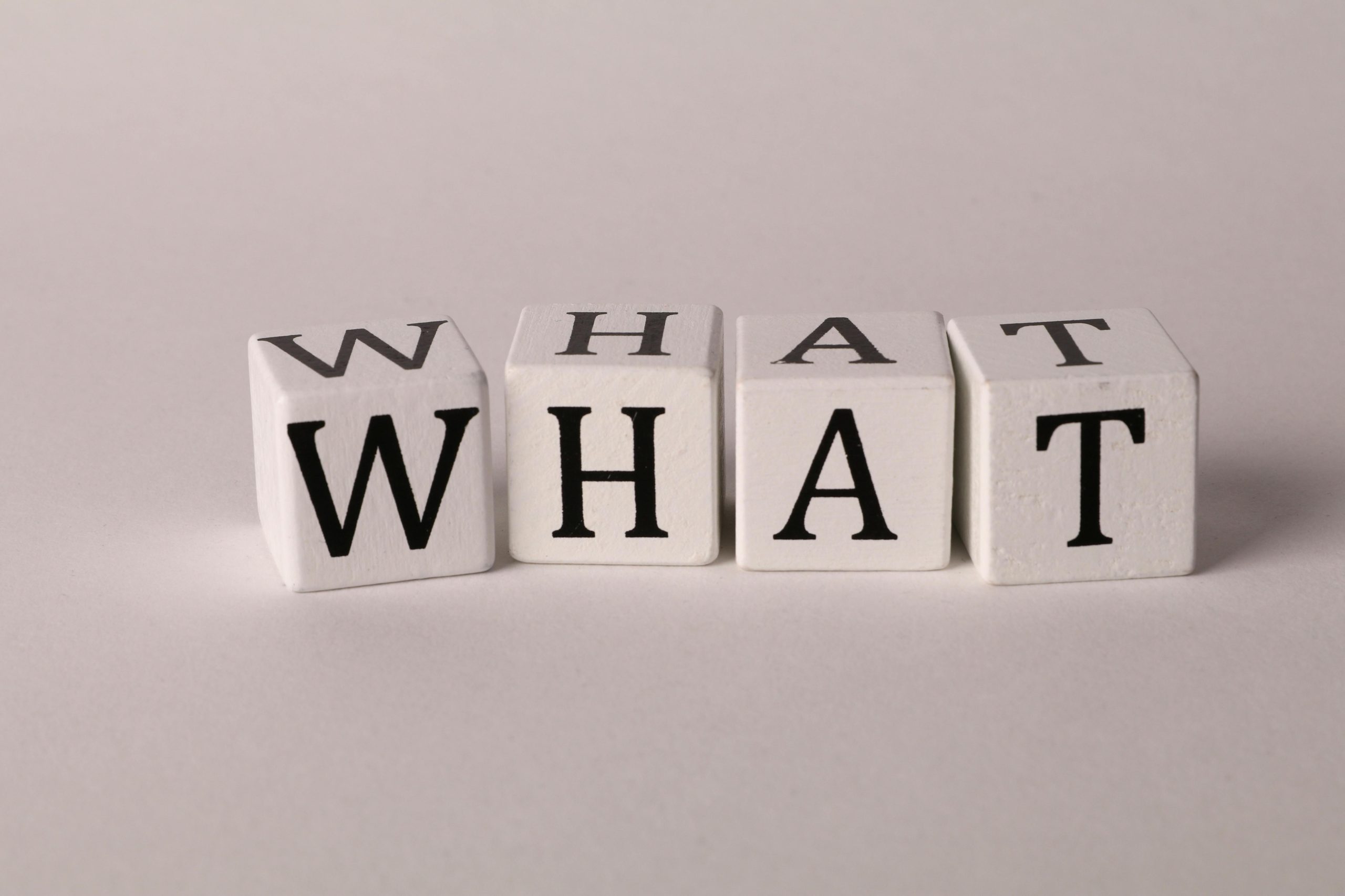 Four white wooden blocks on a light background spelling out the word "WHAT" in bold black letters, symbolizing curiosity or a frequently asked question.