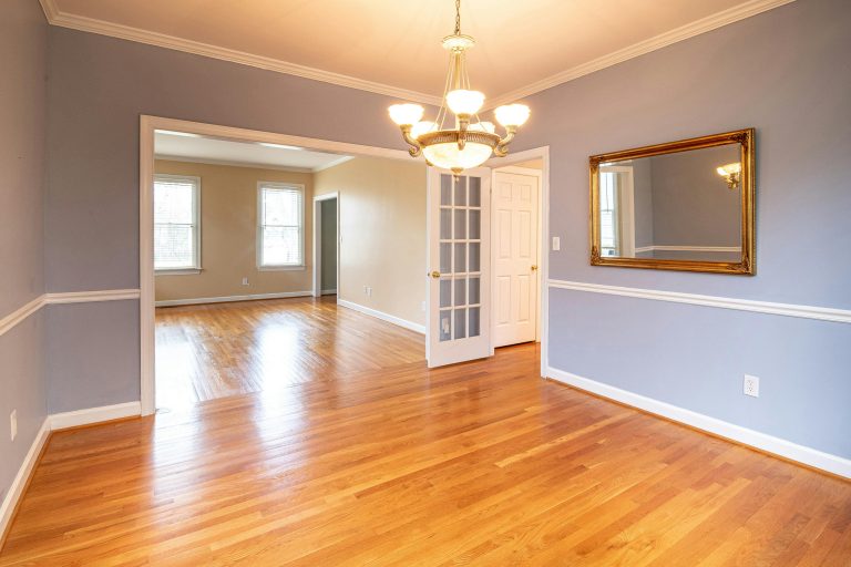 Bright empty dining room with hardwood floors, a gold-framed mirror, chandelier lighting, and a view into an adjacent living space, capturing the quiet stillness of a home ready for its next chapter.