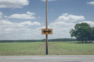 A yellow road sign with black arrows pointing in opposite directions stands on a pole at the edge of a rural field under a blue sky with scattered clouds.