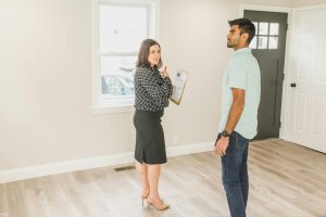 Home buyer standing in the living room looking around intently along with a real estate agent looking at the buyer while holding a clip board