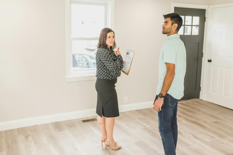Home buyer standing in the living room looking around intently along with a real estate agent looking at the buyer while holding a clip board