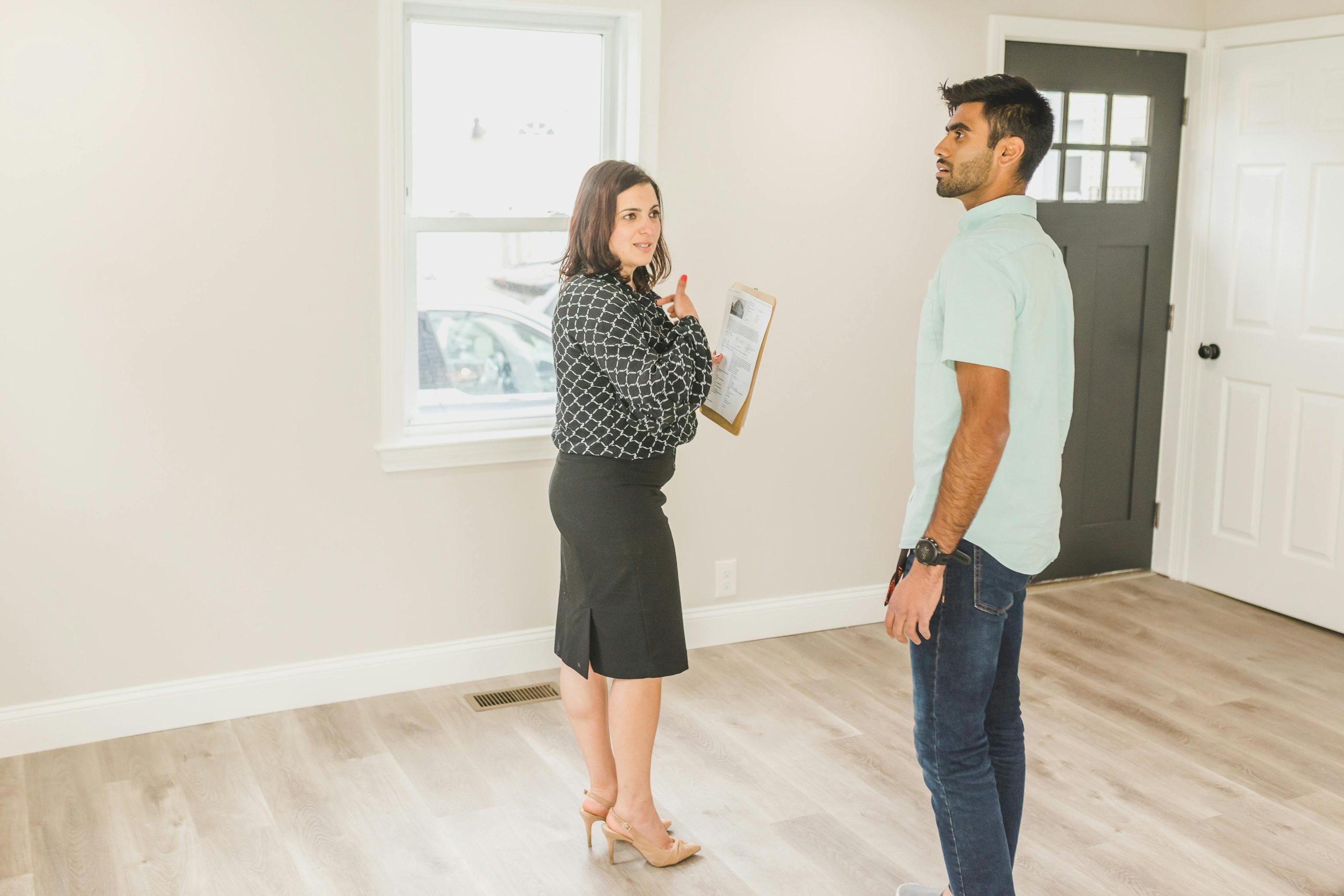 Home buyer standing in the living room looking around intently along with a real estate agent looking at the buyer while holding a clip board