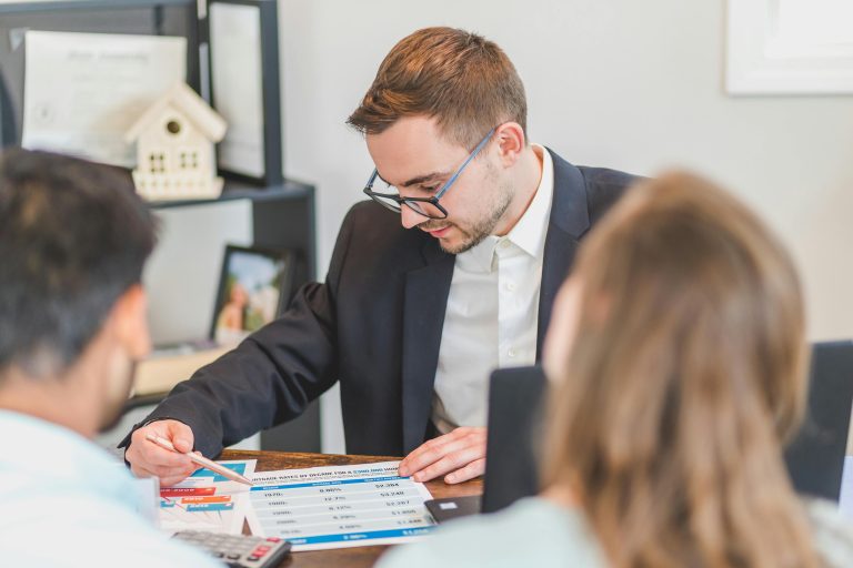 A loan officer in a suit explains mortgage interest rate options to a couple using a printed chart, highlighting the concept of buying down a mortgage rate during the home buying process.