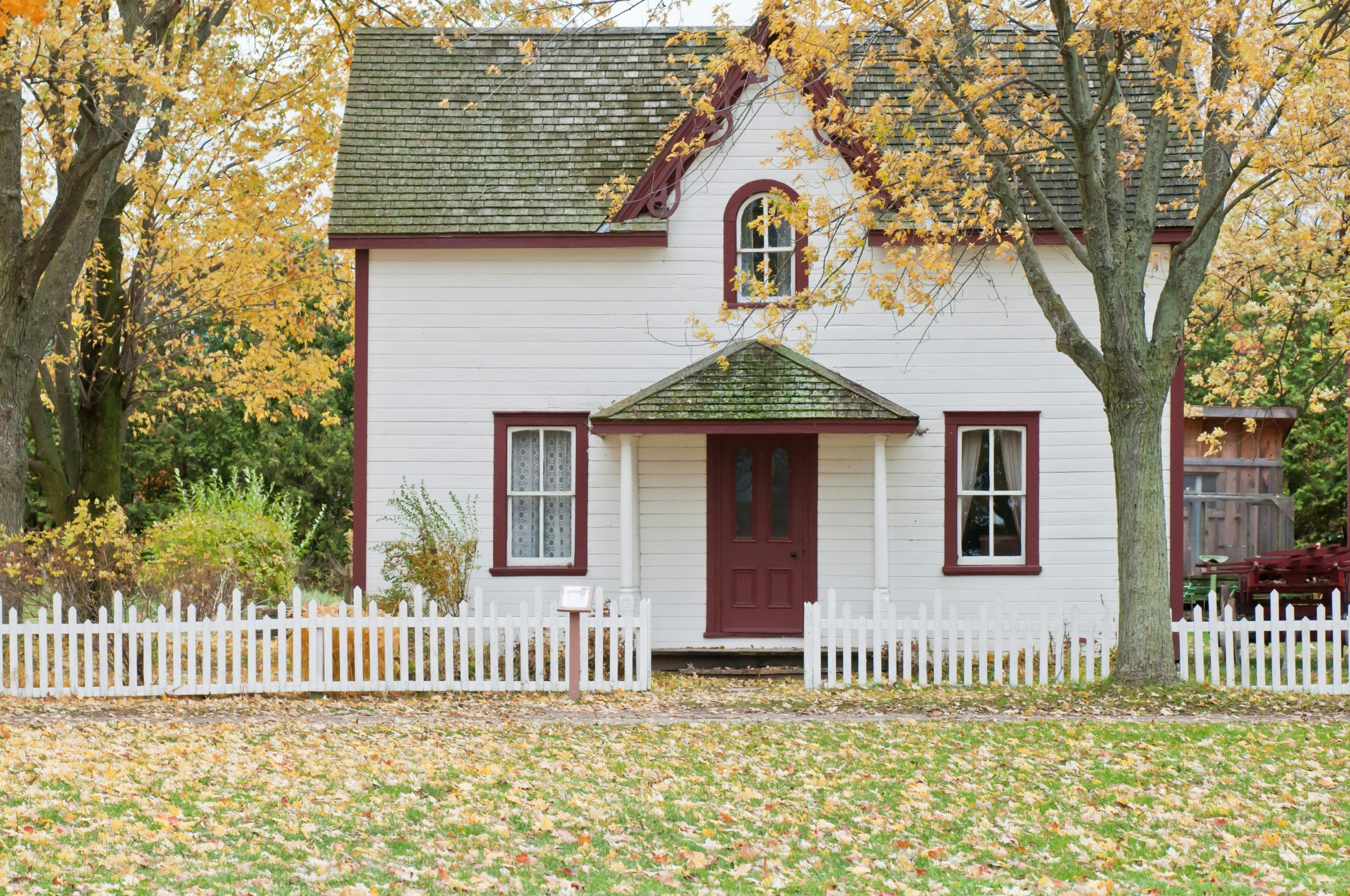 A small white starter home with red trim and a picket fence, surrounded by autumn trees and fallen leaves, representing the concept of affordable starter homes.