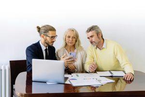 A real estate agent sitting with a couple showing them documents while his computer is open.
