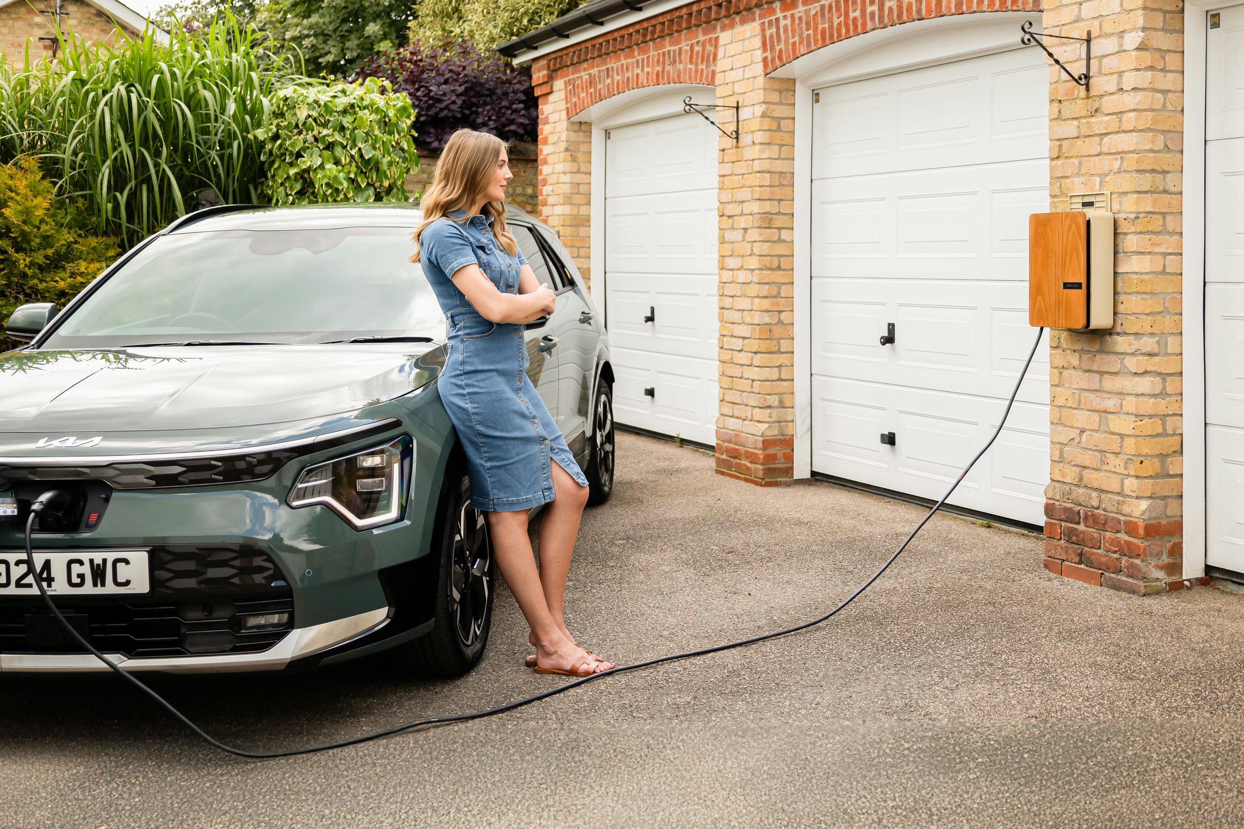 - Unbiased Real Estate Advice: The Truth About Real Estate A woman leans against an electric vehicle charging in front of a brick garage, highlighting the growing trend of EV chargers in residential homes.