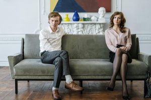 A man and woman sit apart on a gray couch, both looking tense and distant, symbolizing an unmarried couple facing emotional and legal challenges after a breakup while co-owning a home.
