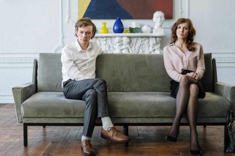 A man and woman sit apart on a gray couch, both looking tense and distant, symbolizing an unmarried couple facing emotional and legal challenges after a breakup while co-owning a home.