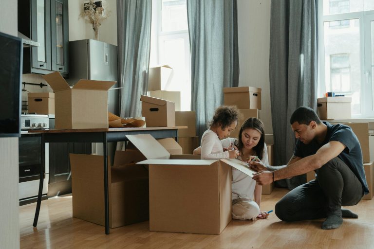 A young family sits on the floor of their kitchen surrounded by moving boxes, writing on paper together as they prepare to move into a new home.