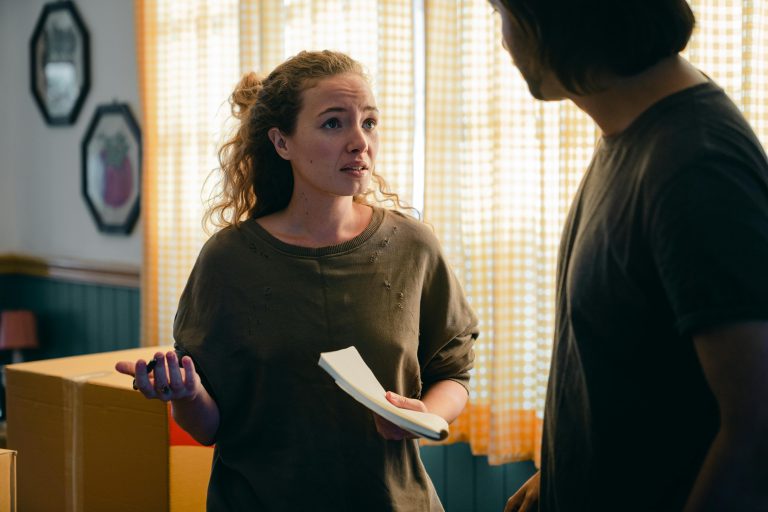 A young couple stands in a room filled with moving boxes, having a serious conversation, with the woman holding a notebook and appearing concerned, reflecting the emotional and uncertain decisions often faced in real estate transactions.