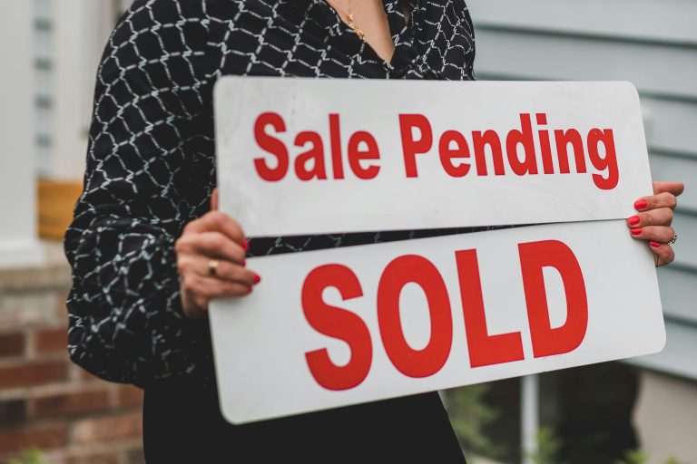 A real estate agent holding a "Sale Pending" and "SOLD" sign in front of a house, symbolizing a home purchase in progress.