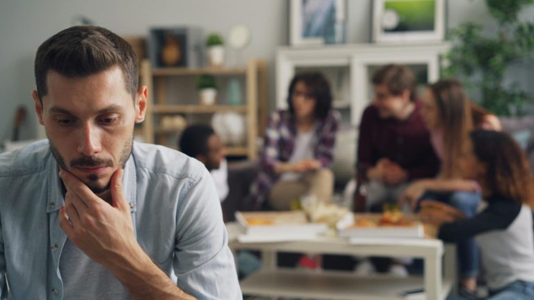 A man sits apart from a group of friends, looking thoughtful and concerned, illustrating the emotional strain that can come from mixing friendships with real estate decisions.