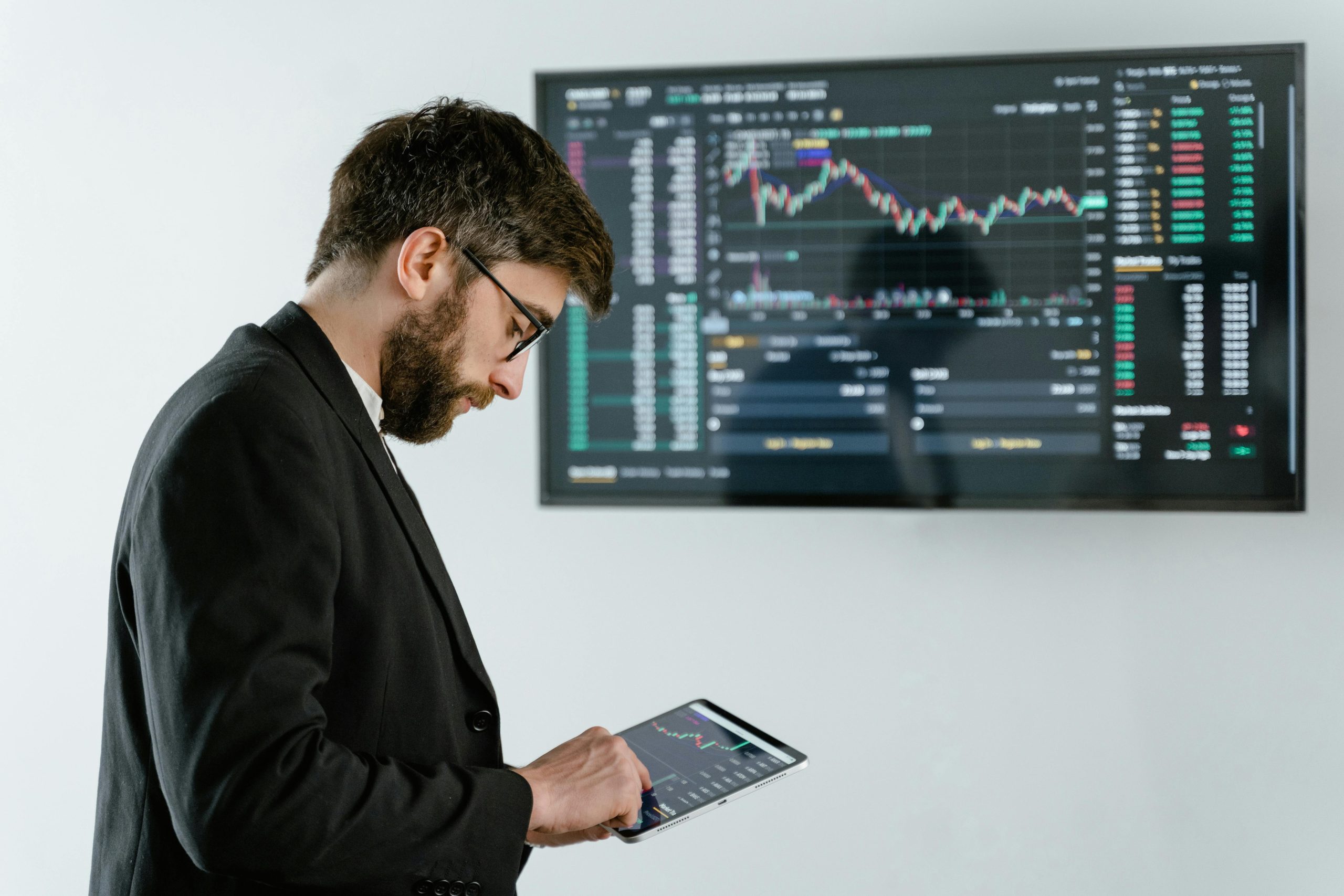 - Unbiased Real Estate Advice: The Truth About Real Estate A man in a suit studies real estate market data on a tablet while a financial graph displays on a large screen in the background, representing the shifting housing market with more sellers than buyers.