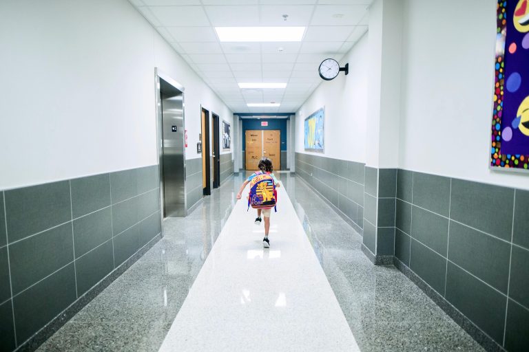 Young child wearing a colorful backpack running down a school hallway toward classroom doors