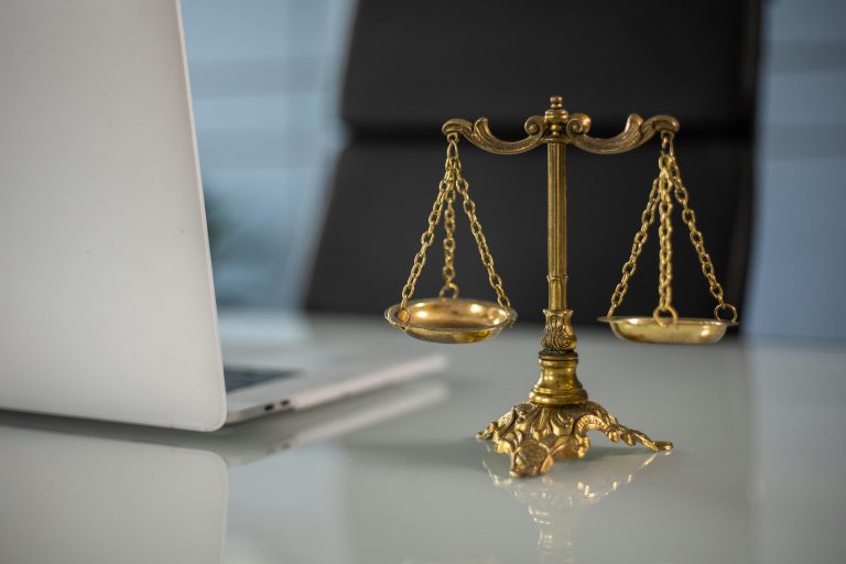 A brass scale of justice next to a laptop on a glass desk, symbolizing the legal support of a real estate attorney in property transactions