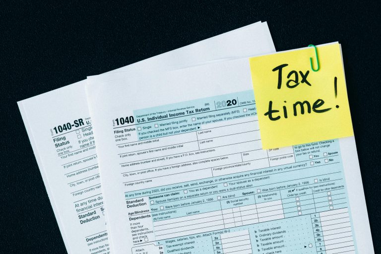 Close-up of U.S. 1040 individual income tax return forms with a yellow sticky note reading "Tax time" clipped to the top of the papers on a desk