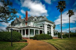 Large white luxury mansion with wraparound porch, tall columns, striped awnings, and manicured landscaping under a blue sky with palm trees