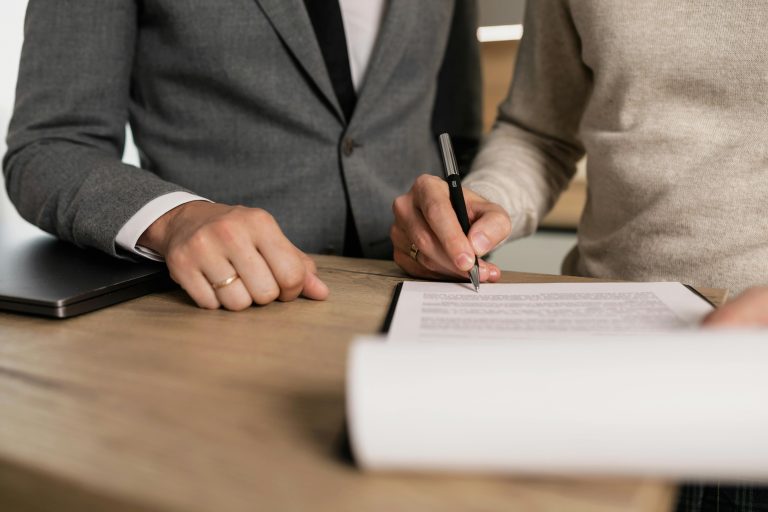 Close-up of two people signing a contract at a wooden desk, one in a gray suit and the other in a beige sweater, with a black pen and a closed laptop nearby.