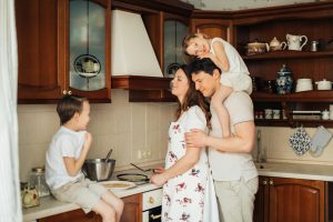 A happy family spending time together in a cozy kitchen. The mother is cooking at the stove while the father stands behind her holding their young daughter on his shoulders. Their son sits on the countertop, smiling and chatting with them.