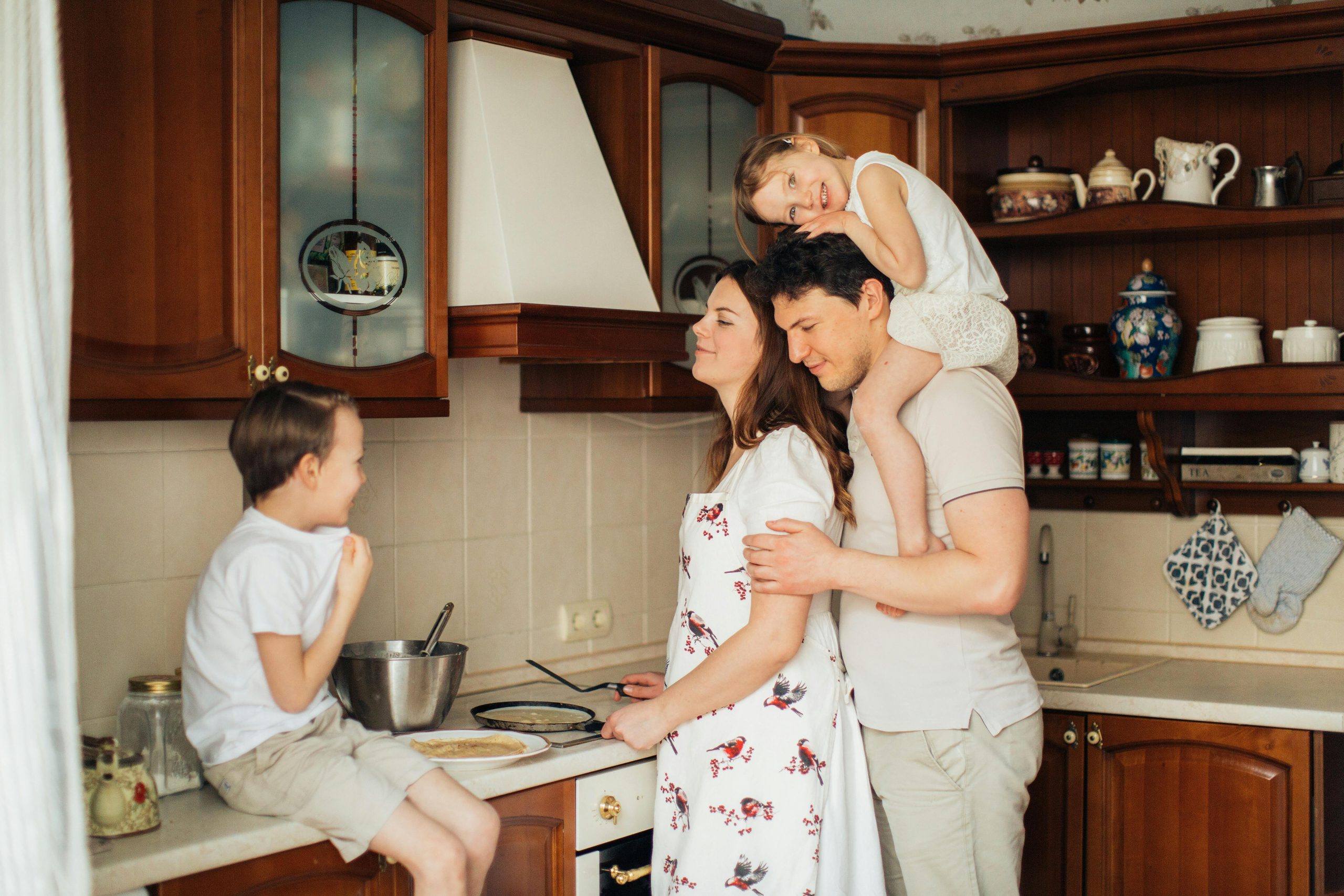- Unbiased Real Estate Advice: The Truth About Real Estate A happy family spending time together in a cozy kitchen. The mother is cooking at the stove while the father stands behind her holding their young daughter on his shoulders. Their son sits on the countertop, smiling and chatting with them.