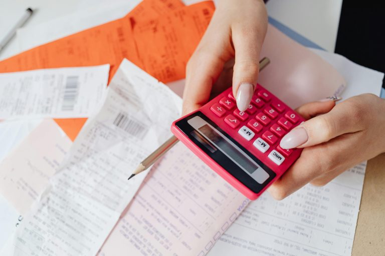 Person holding a pink calculator and pencil while reviewing receipts and financial documents, calculating expenses, budgeting, money management, seller concessions, home selling costs, financial planning, real estate transaction costs