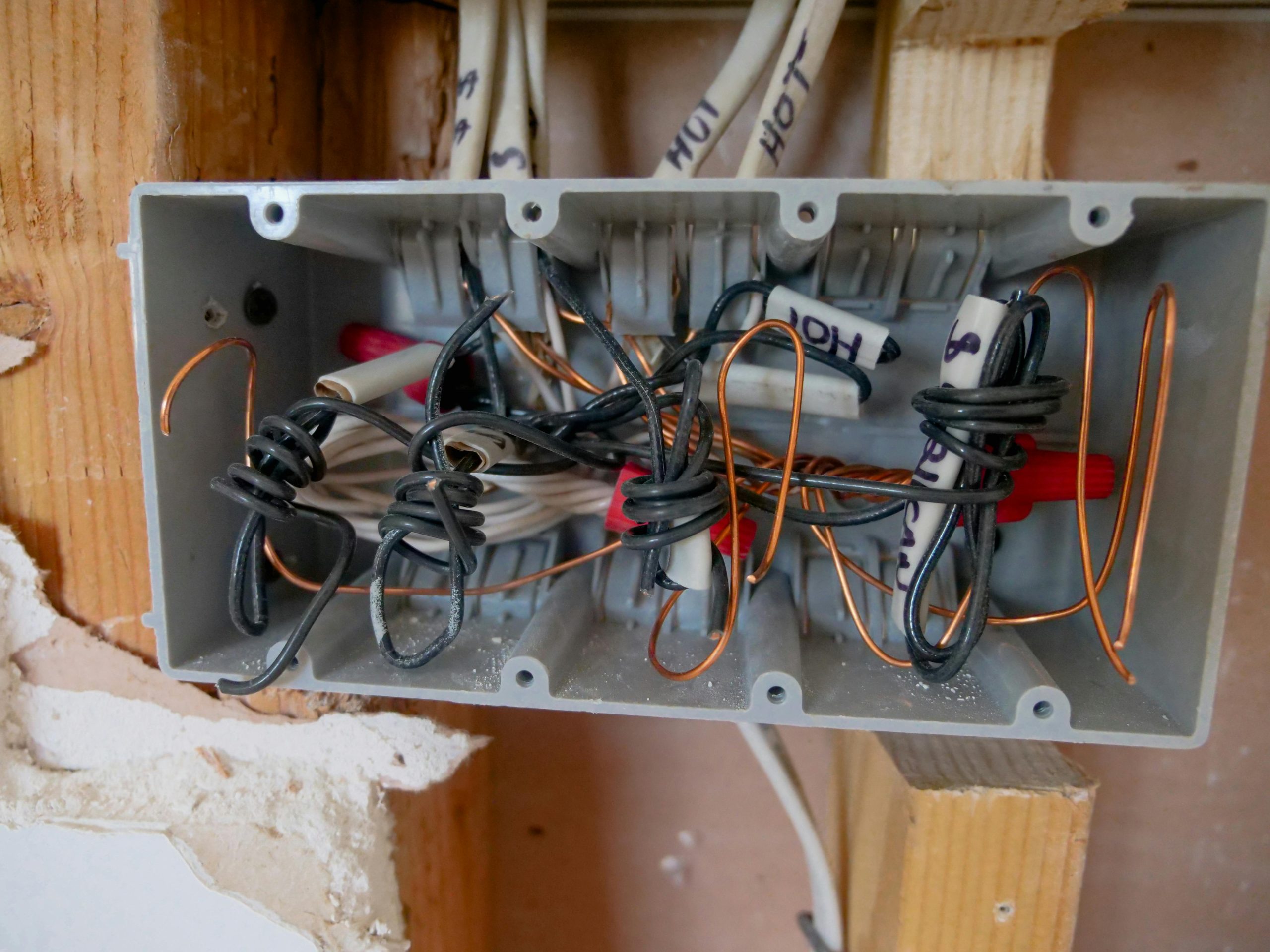 Close-up view of an open electrical junction box with exposed copper and black wires, wire nuts, and labeled cables, showing ongoing electrical work inside a wall