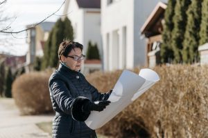 Woman standing outdoors in a residential neighborhood, wearing a dark quilted coat and gloves, looking at and holding large rolled architectural blueprints, with houses and shrubs in the background, winter season.