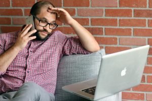A frustrated man wearing glasses and a red checkered shirt sits on a couch while talking on the phone and looking at a laptop, appearing concerned or stressed.