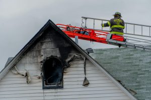 A firefighter on a ladder inspecting a burned house roof and window after a fire, showing the risks of being underinsured as a homeowner.