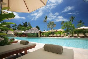 Luxury resort pool with lounge chairs, umbrellas, and palm trees under a bright blue sky, symbolizing a real estate agent taking a vacation.