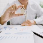 Person holding a small house keychain with a key over printed financial charts and a laptop, representing analysis of a rental property investment.