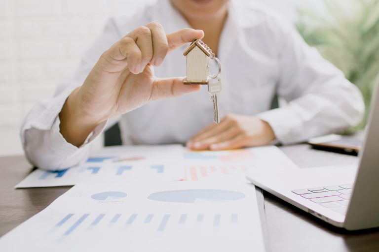 Person holding a small house keychain with a key over printed financial charts and a laptop, representing analysis of a rental property investment.