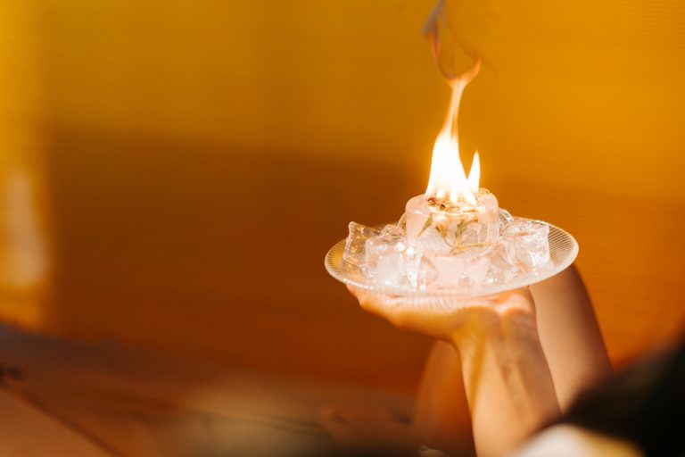 Hand holding a small plate of ice cubes with a flame on top against a warm yellow background, symbolizing hot and cold conditions in the housing market.
