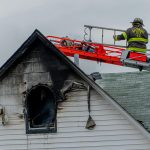 Firefighter on an aerial ladder beside a house with smoke and charred siding after a fire, illustrating the risks a last minute clue report can reveal before closing.