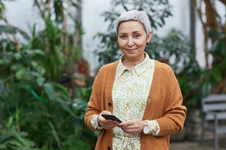 Smiling older American woman holding a smartphone in a garden setting, representing modern housing and lifestyle options for seniors.
