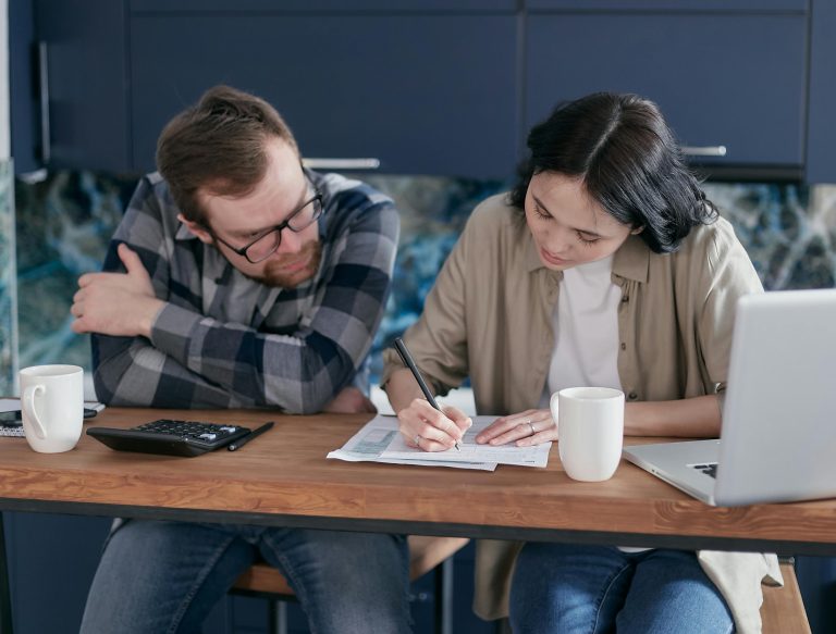Couple at a kitchen table reviewing seller disclosure paperwork with a calculator, coffee mugs, and a laptop, preparing accurate documents before listing their home.