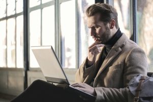 Thoughtful man in a camel coat working on a laptop by large windows, researching a home that is back on the market.