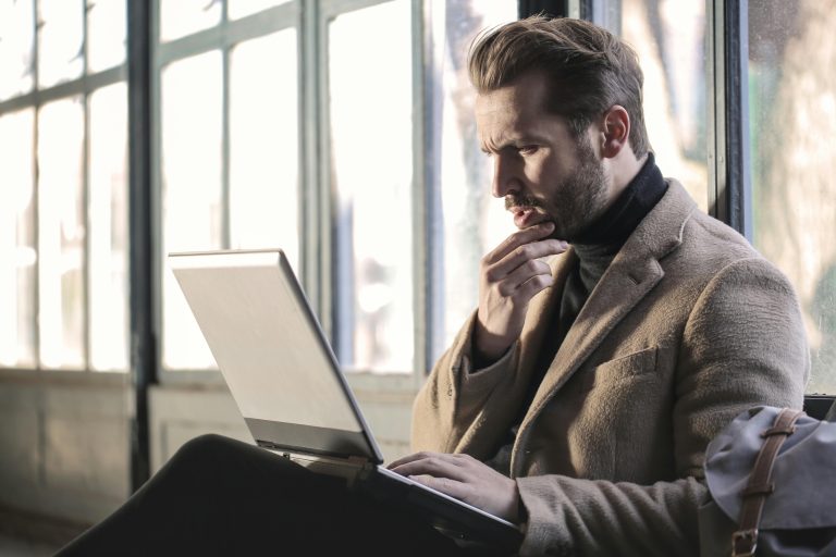 Thoughtful man in a camel coat working on a laptop by large windows, researching a home that is back on the market.