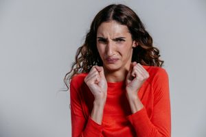 Woman in a red sweater grimacing and pulling her hands close to her face, reacting to an unpleasant smell on a neutral background.