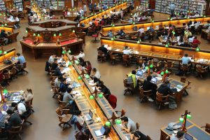 Overhead view of a busy university library with many college student learners studying at long wooden tables.
