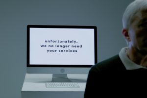 Close up of a desktop screen that reads “unfortunately, we no longer need your services” with a worried person slightly out of focus in the foreground.