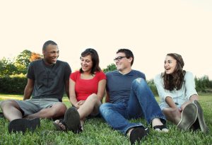 Four young adults sitting together on grass, smiling and talking, representing young buyers considering homeownership decisions.